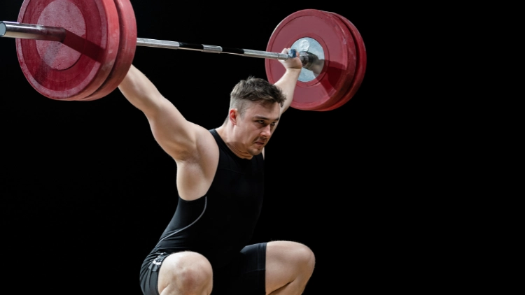 A guy in a black tank top and with short hair who is holding 275 above his head after doing a clean and press with a snatch grip. 