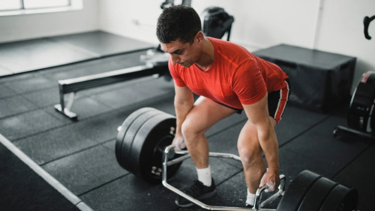 A man wearing a red activewear shirt, black short, and shoes with white socks pulling up a hex bar to perform a deadlift in a gym with black platform where he is standing. 