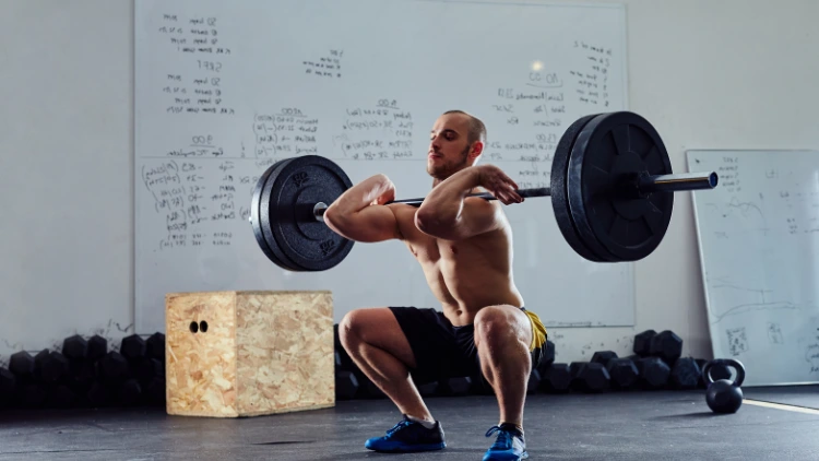 A topless man wearing black shorts and blue shoes is performing a barbell front squat using a barbell with two weighted plates in a studio with a plyo box and dumbbells on the floor and a white board with bunch of writings on the wall in the background.