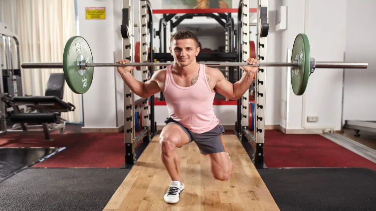 A man in pink tank top, grey shorts and white shoes performing barbell lunges using a barbell with green plates, in a gym with various equipment in the background.