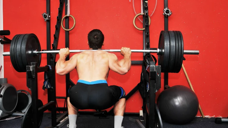 A topless man in black shorts and white socks is doing barbell back squats in a commercial gym with red wall and fitness ball on the floor in the background.