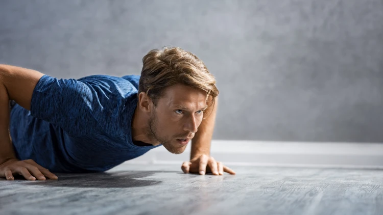 A blonde man in a blue shirt with his body extended in a plank position and his hands are supporting his weights on the ground doing a push-up on a grey and shiny concrete floor.