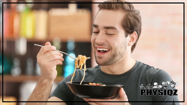 A man wearing a black shirt eating a pasta on a black bowl using a fork in a room with unidentifiable objects in the background.