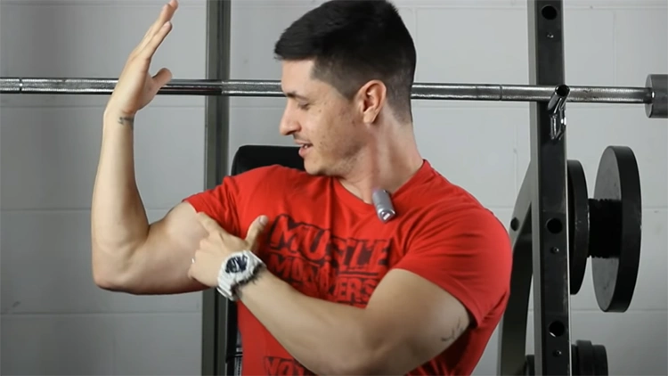A man in a red shirt and with a white wrist watch on his left arm is pointing to his right bicep with a silver barbell behind him. 