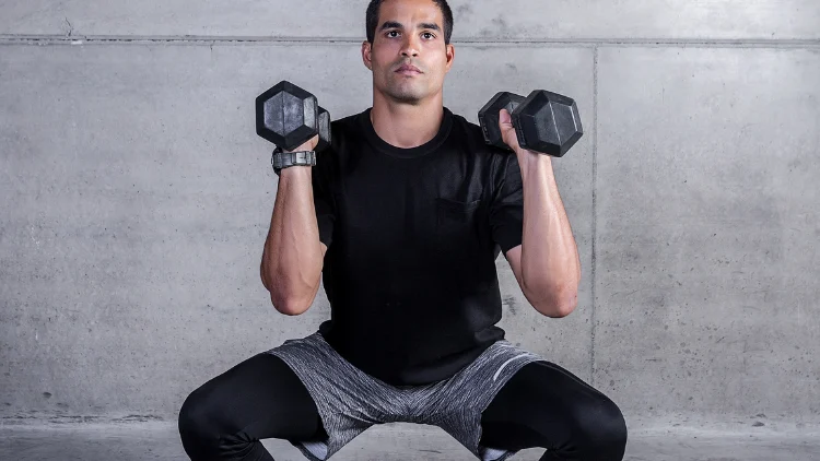 A man in black shirt, gray shorts, and black tights and wearing a watch is performing a Dumbbell Squat Clean exercise, the dumbbell is in his shoulder in a concrete background.