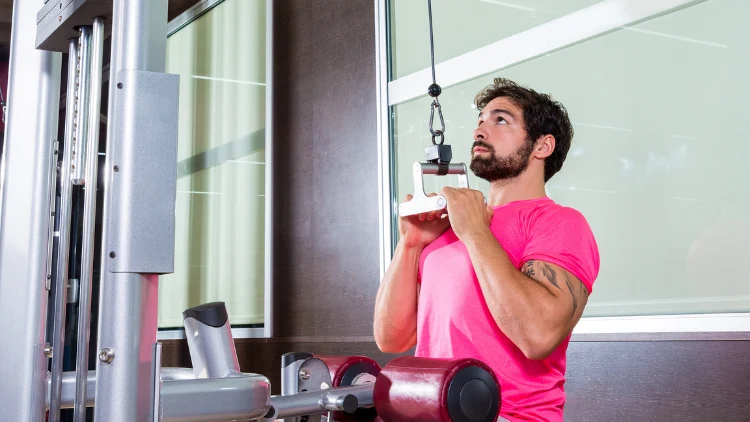 A bearded man wearing a pink t-shirt is performing a close grip lat pulldown exercise in a gym with window behind him in the background.