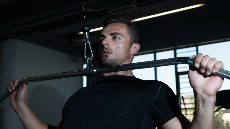 A bearded man wearing a black activewear shirt is focused in performing a wide grip lat pulldown exercise in a dimly lit studio with large glass window in the background.