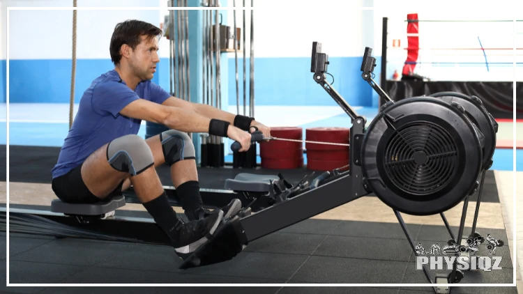 A guy in a blue shirt has on black wrist bands and grey knee sleeves on as he pulls on a rowing machine. 