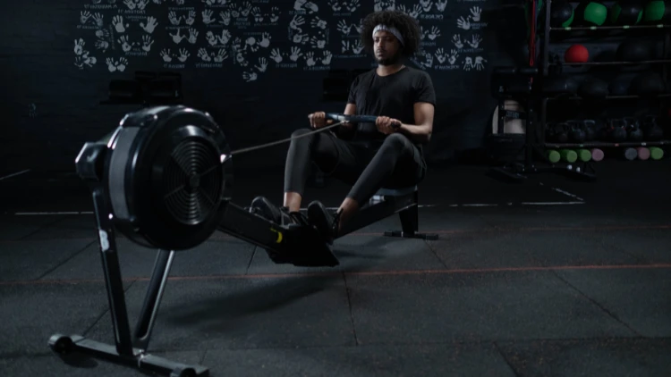 A man with an afro, wearing a black shirt and black pants, is using a rowing machine in a studio that has handprints on the walls and various gym equipment visible in the background.