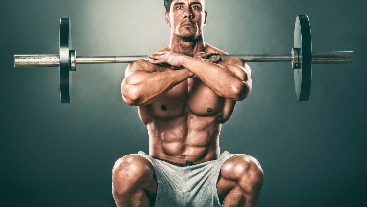 A topless muscular man wearing a grey short is performing a front squat cross grips with a barbell in a studio with green wall.