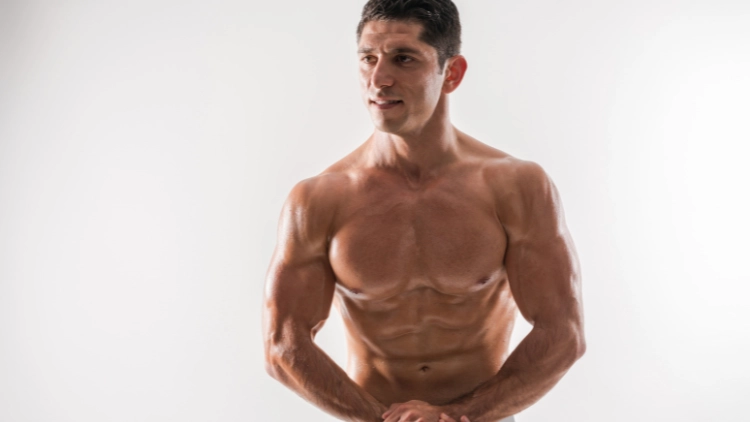 A white man posing in a studio that shows he has a six-pack, striated shoulders, and low bicep insertions from his large bicep belly. 