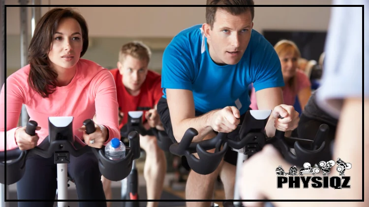 A woman in a pink top and black pants are wondering the amount of Peloton calories burned, and a man in a blue top and black shorts, are riding stationary bikes in a gym while observing a demonstrator, and in the background, a man in a red shirt and black shorts, and a woman in a pink top can be seen.