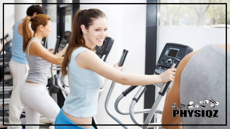 A pale women in a light blue shirt is holding the cardio machine handles that monitor heart rate and calories burned and there's another woman in white pants and a grey top behind her. 