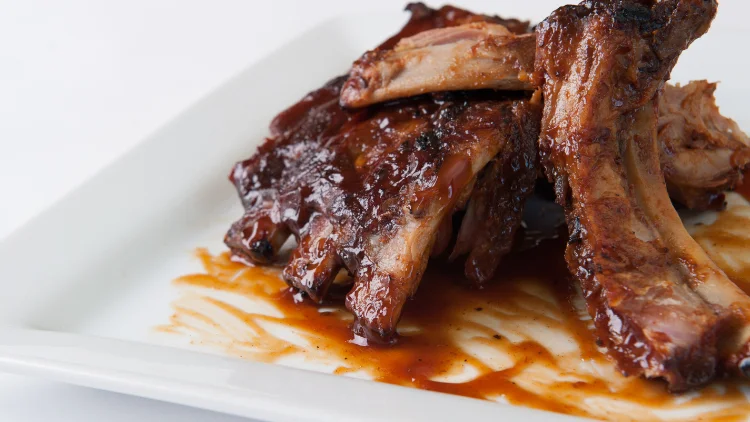 On a white plate is a serving of baby back ribs glazed in savory sauce and displayed on a white surface.