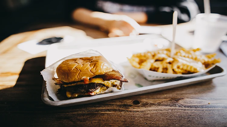 On a silver tray, a cheeseburger and a serving of chips with a spoon on it is displayed on a wooden table with a person blurred in the background.