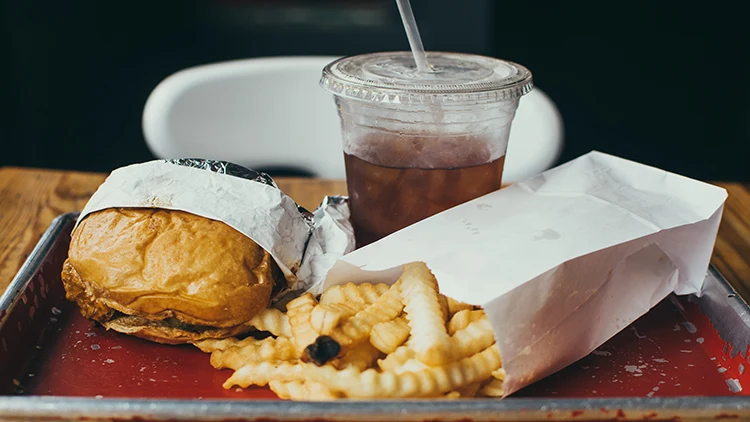 A meal composed of a burger half-wrapped in an aluminum foil, fries in a white paper bag, and iced tea in a clear plastic cup with straw served on a silver tray with red bottom and placed on the wooden table in a restaurant.
