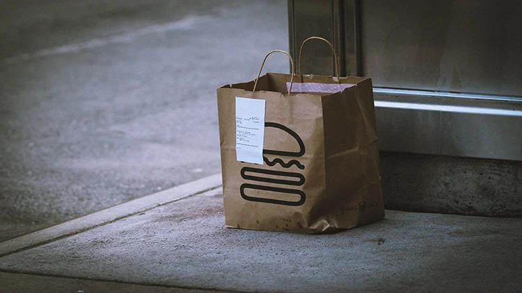 A brown paper take-out bag with a receipt attached and a burger logo, a branding of the Shake Shack, placed on a pavement outdoors.