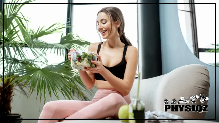 A blonde haired woman is sitting on a love cushion inside a living room with blue walls and a house plant as she eats a salad while wearing a black tank top and light pink pants. 