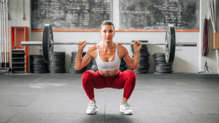 A fit woman wearing a white tank top, red pants and white shoes is performing a barbell back squat using a barbell with only one weighted plate in a studio with weighted plates on the floor and a chalkboard filled with writings in the background.