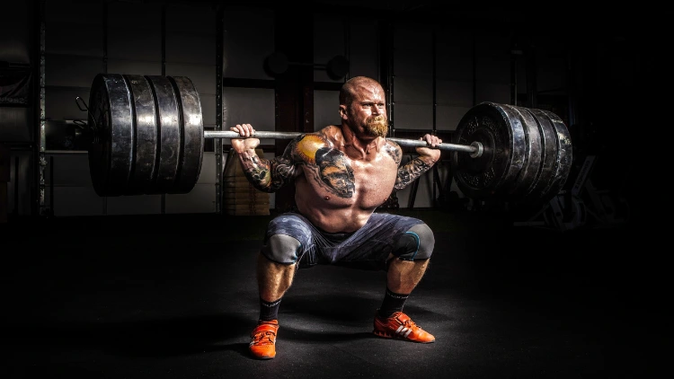 A topless muscular man with tattoos on his chest, and both arms, wearing a blue pants and orange shoes is doing a barbell back squat with 4 weighted plates inside a dimly lit gym.