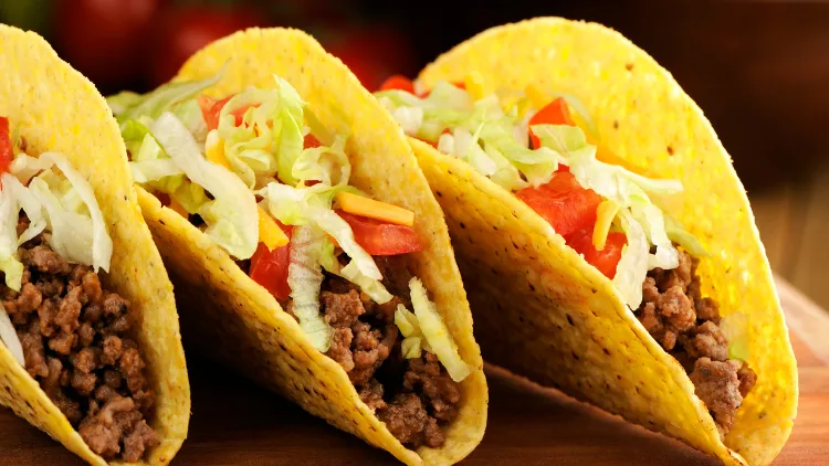 Three crunchy tacos on a plate, each taco shell is filled with seasoned ground beef, shredded lettuce, diced tomatoes, and shredded cheese.