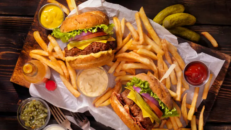 On a wooden tray are servings of two burgers made of beef patty, lettuce, tomato, onions, and cheese, and surrounding it are fries, and dipping sauces such as mustard, ketchup and chili.