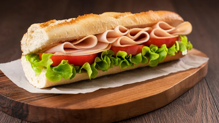 A sandwich snack made of turkey meat, cheese, tomato and vegetable salad with a paper wrapper below it and served on a wooden board which is then placed on top of a wooden surface.