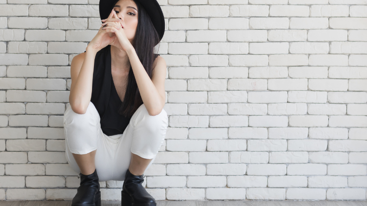 Asian woman wearing a black hat, black top, and white pants, with black shoes, sitting in an Asian squat position, her elbows are resting on her knees, and her arms are crossed in front of her mouth, she appears to be in a contemplative or reflective state, with a serene expression on her face.