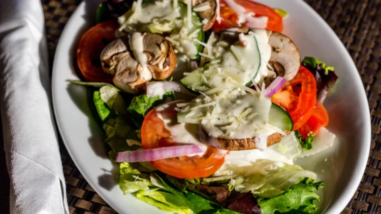 A plate of fresh vegetable salad, drizzled with a creamy ranch dressing, the salad includes a colorful assortment of vegetables such as lettuce, carrots, cucumbers, tomatoes, and red onions, all sliced or chopped into bite-sized pieces.