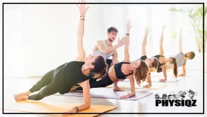 Four women and one man are doing a wall walk alternative or the side plank on yoga mats inside a well lit room that has a green plant and white vase in the corner and there's also an instructor coaching them through the movement.