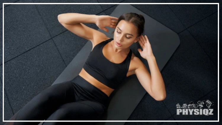 A woman wearing black tank top and black pants doing ab workout on a black yoga mat in a studio with black tiled floors.
