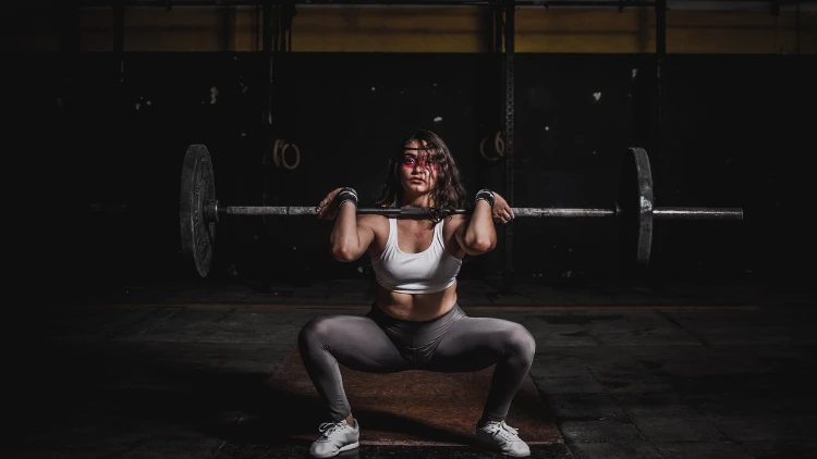 A woman wearing a white top and gray pants, performing a squat exercise with a barbell, she is lifting the barbell up to her shoulders while in a squat position, engaging her leg and core muscles.