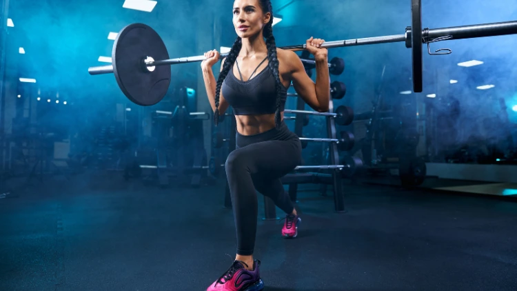 A woman with braided twin tails, wearing a black sports bra and leggings, and pink shoes is performing a barbell lunges in a foggy and dimly lit gym with barbell rack visible in the background.