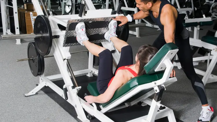 A woman wearing a red tank top, black pants, and white shoes is doing an exercise using a leg press machine with the help of a fitness instructor beside her, wearing a black tank top, pants and white shoes in a gym with concrete floor.