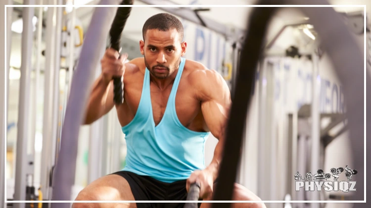 A black man working out 7 days a week is wearing a sky-blue tank top and black shorts while using black battle ropes on day 2 of his program and inside a gym with a pull up bar in the background.