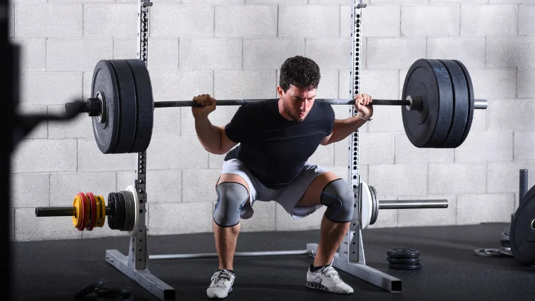 A young man wearing a black activewear shirt, gray short and knee guards, and white shoes is performing a heavy weight barbell back squats using a barbell with two black weighted plates in a gym with concrete brick walling and black floor.