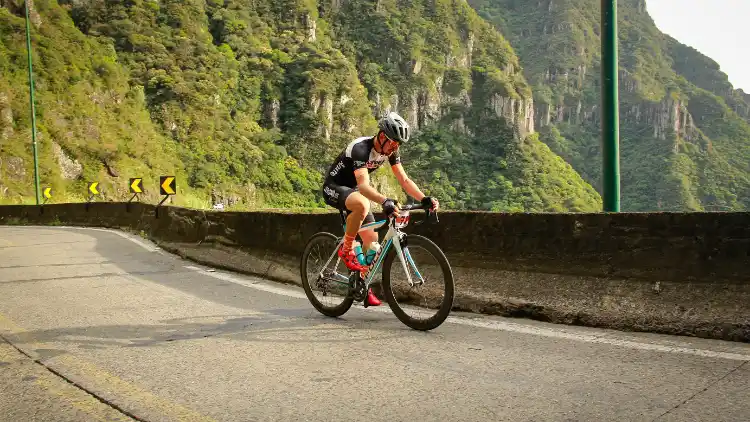 A man wearing a black and white cycling suit, red shoes with orange socks, and a black helmet is riding a bike on a road with arrow signs, while a cliff can be seen in the background.