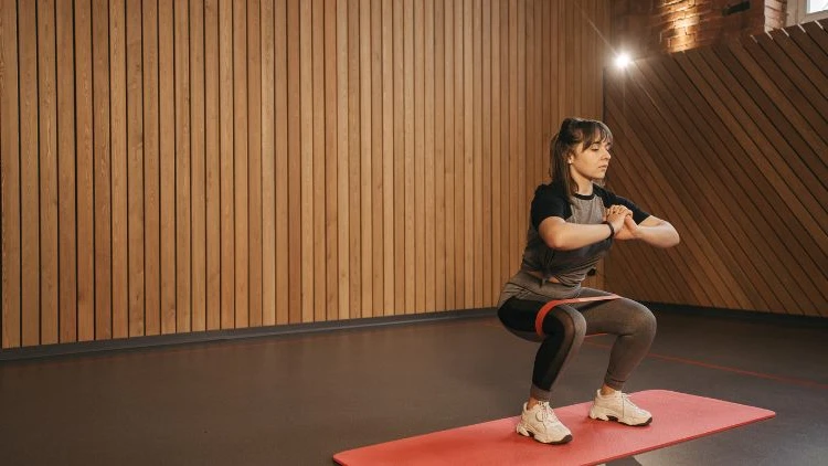 A woman wearing a matching black and grey top and pants doing a squat with a resistance band on a red yoga mat in a studio with black floor and vertical wood slat walling, and a light can be seen in the background.