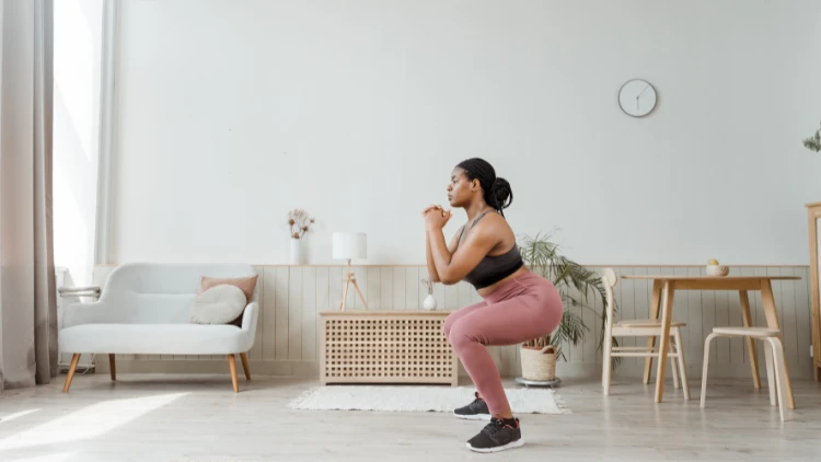 A woman wearing black tank top, pink pants, and black shoes doing squats in a room with various furniture in the background.