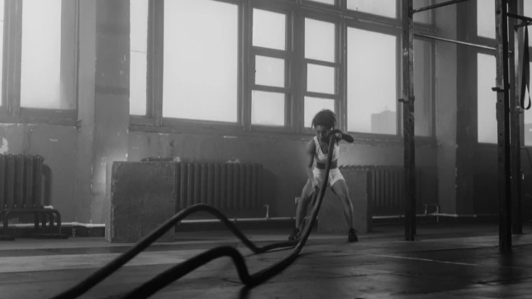 A black and white picture of a woman wearing a white tank top, white shorts and black shoes doing a battle rope exercise in a studio with large windows in the background.