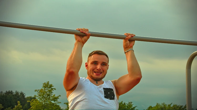 A man wearing white tank top and a bracelet doing a pull ups exercise outdoor with trees in the background.