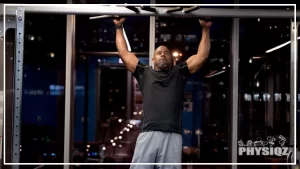 A middle aged black man who is muscular and bald is wondering the average pull ups by age as he hands from a wide grip pull up bar and he's wearing a back shirt, and grey shorts and inside a commercial gym with a mirror in the background.