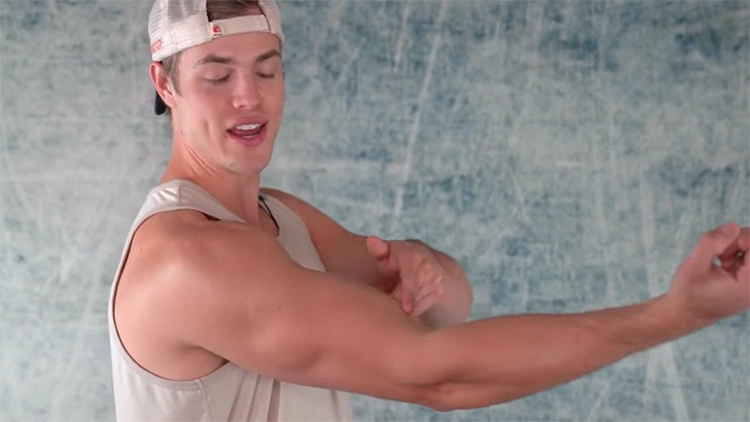 In a room with blue and white walls, a guy wearing a white snapback and a tan tank top is pointing to the insertion point of his right biceps, questioning whether his arm genetics are really bad or if it's just a problem with his training.