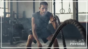 A man wearing black t-shirt with both sleeves rolled up and black shorts is using all of the battle rope muscles worked as he does alternating waves inside a dark gym with a pair of white gymnastics rings hanging in front of a window and black rubber weight plates laying on the ground. and black shorts doing an exercise using a battle rope in a gym.