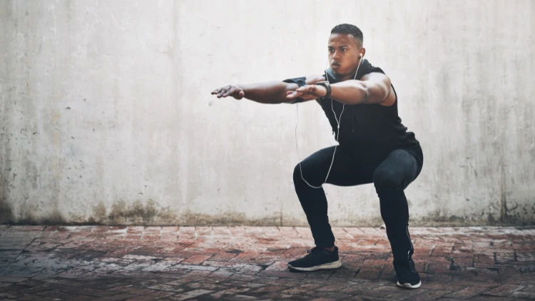 A muscular Black man wearing a black tank top and pants and earphones is performing body weight squats outdoors on a concrete surface, he has a focused expression on his face, and his arms are extended forward for balance.