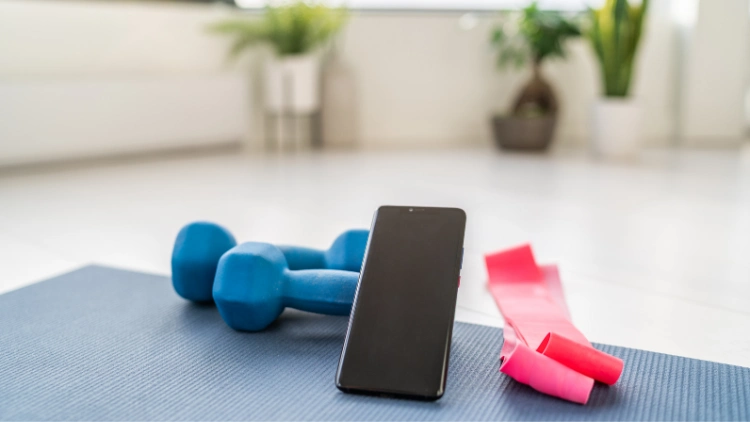 A phone leaning to a blue dumbbell with a resistance band beside the phone placed on gray yoga mat with blur plants in the background.