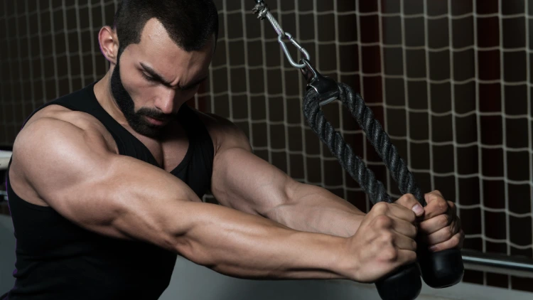 Bearded white man in black tank top is performing a overhead cable triceps extension exercise with square net seen in the background.