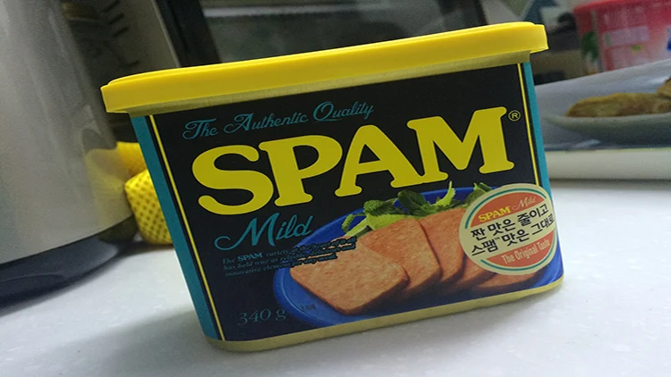 A yellow and blue can of spam in mild variation displayed on a white countertop beside kitchen appliances.