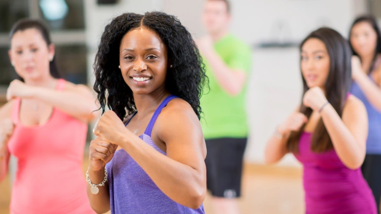 A woman with tan skin and curly hair, wearing a purple tank top is leading a cardio workout class with other people blurred in the background wearing colorful tank tops such as pink, blue, violet and green.