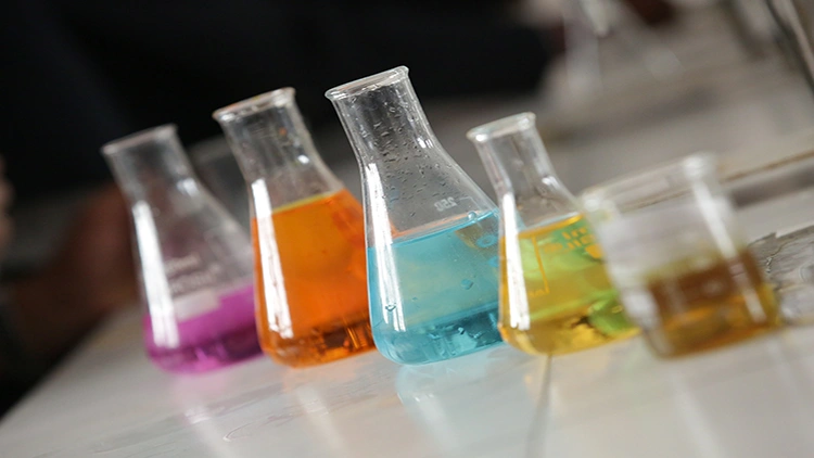 Five chemistry laboratory flasks of different sizes, filled with liquids of various colors such as purple, orange, blue, yellow, and golden brown, are arranged in order of height on a laboratory countertop.
